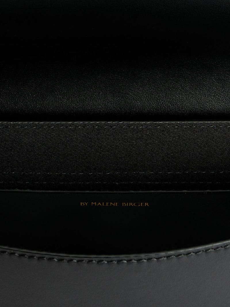 By Malene Birger Maellon veske i blankt, sort kuskinn, med diskret monogram i front og justerbar stropp.

