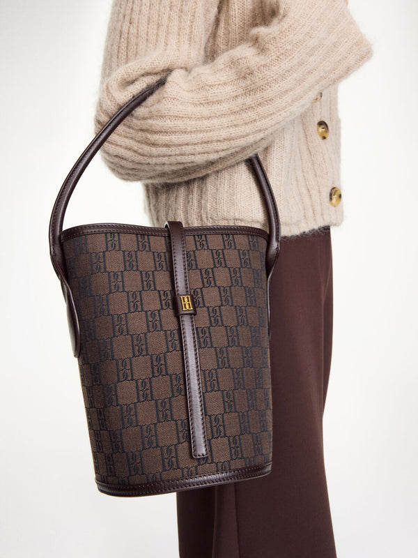 By Malene Birger Maryllon veske i brun, med bøtteform, laget av resirkulert bomull og polyester, og et gullfarget monogram-emblem i front.