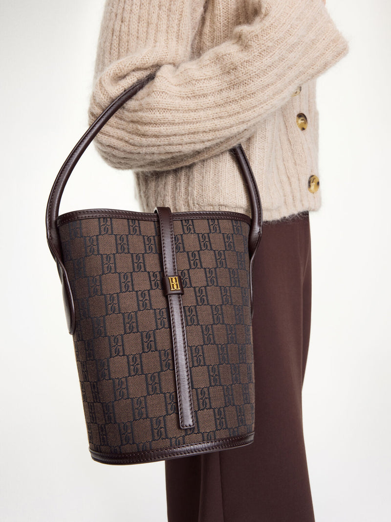 By Malene Birger Maryllon veske i brun, med bøtteform, laget av resirkulert bomull og polyester, og et gullfarget monogram-emblem i front.
