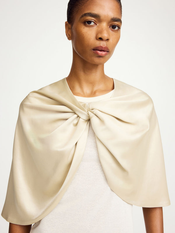 By Malene Birger Pinlos cape i kremfarget sateng, laget av en bærekraftig blanding av acetat og polyester, med knutedetaljer i front.
