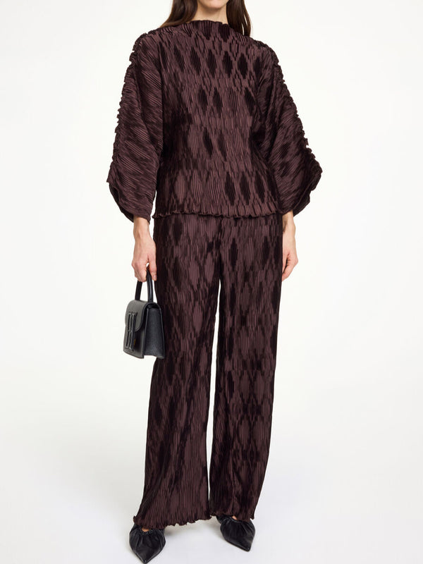 By Malene Birger Tralluman bluse i mørkebrun, laget av 100% resirkulert polyester, med plissé struktur, båtutringning og vide ermer.