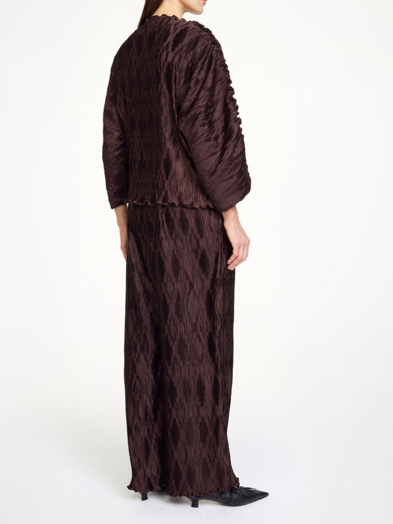 By Malene Birger Tralluman bluse i mørkebrun, laget av 100% resirkulert polyester, med plissé struktur, båtutringning og vide ermer.