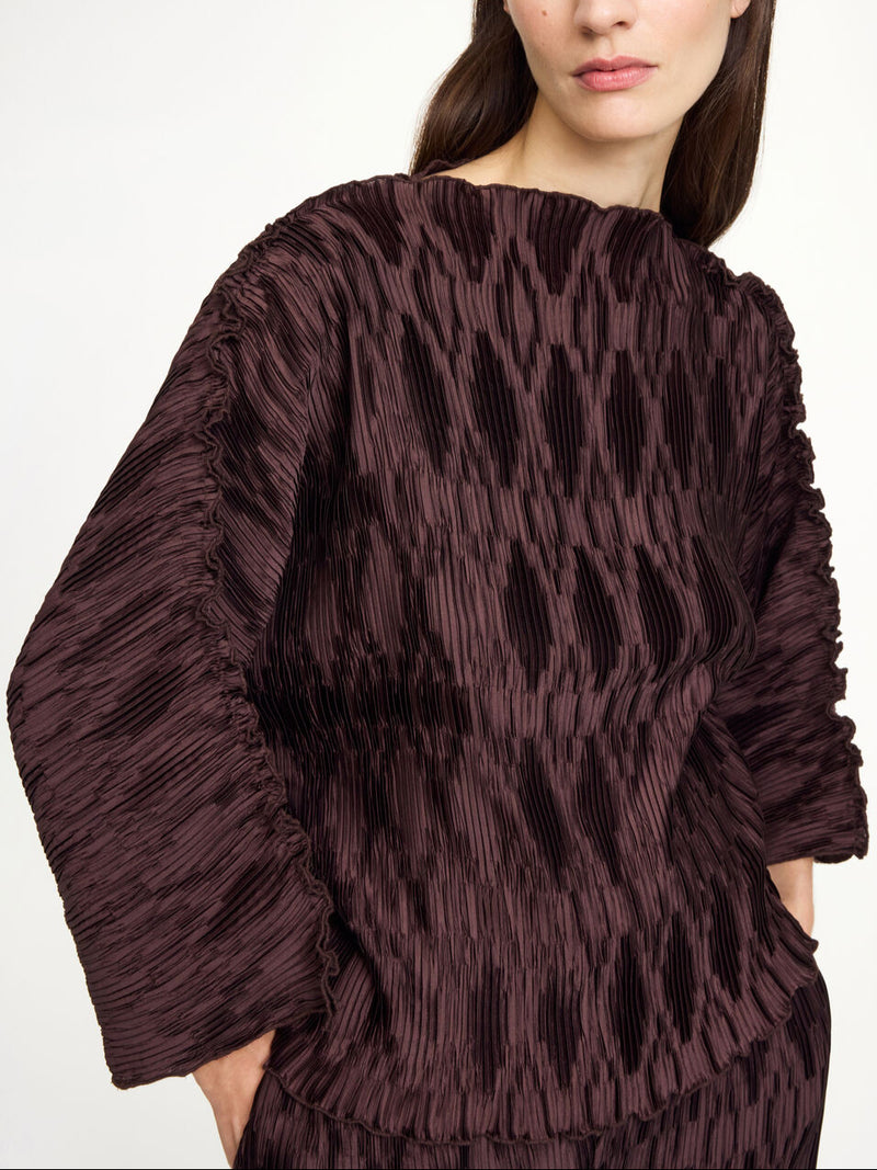 By Malene Birger Tralluman bluse i mørkebrun, laget av 100% resirkulert polyester, med plissé struktur, båtutringning og vide ermer.